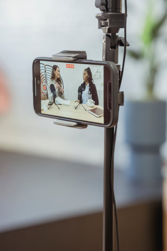 Young diverse women sitting at table with microphones and gadgets and talking while recording podcast on smartphone placed on tripod in studio